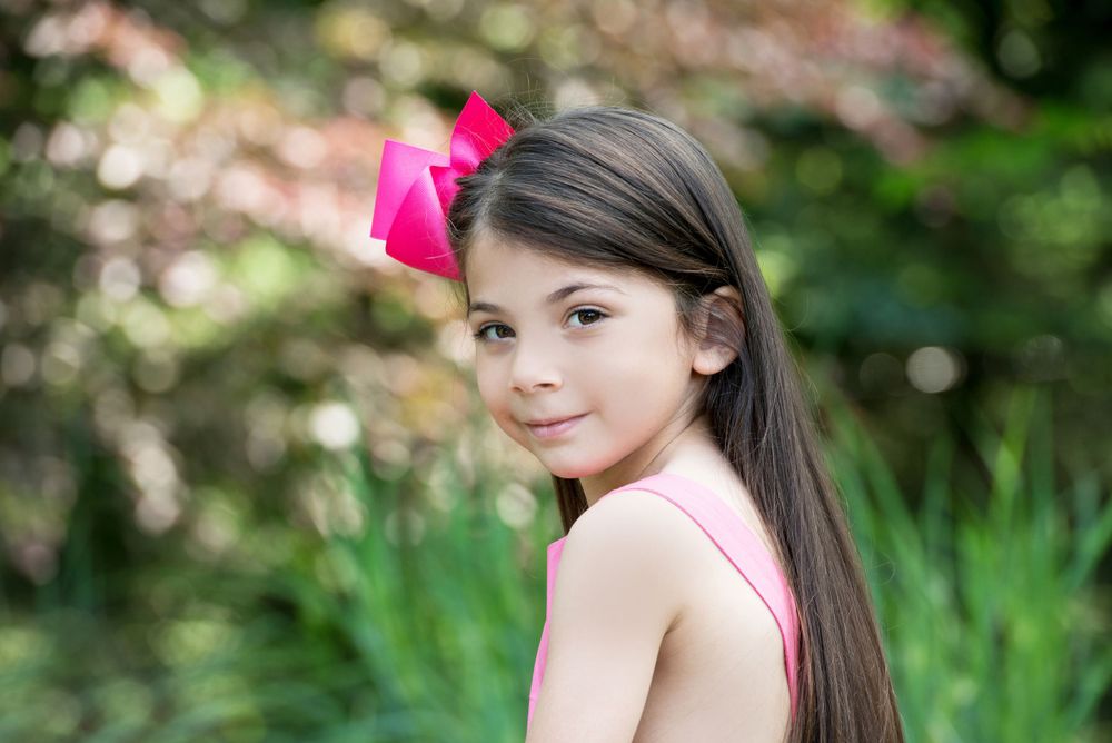 Outdoor portrait with pink bow accessory against blurred garden background.