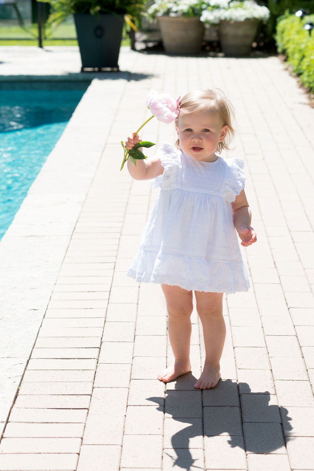 Small child in white dress holding flower while walking beside pool on sunny day.