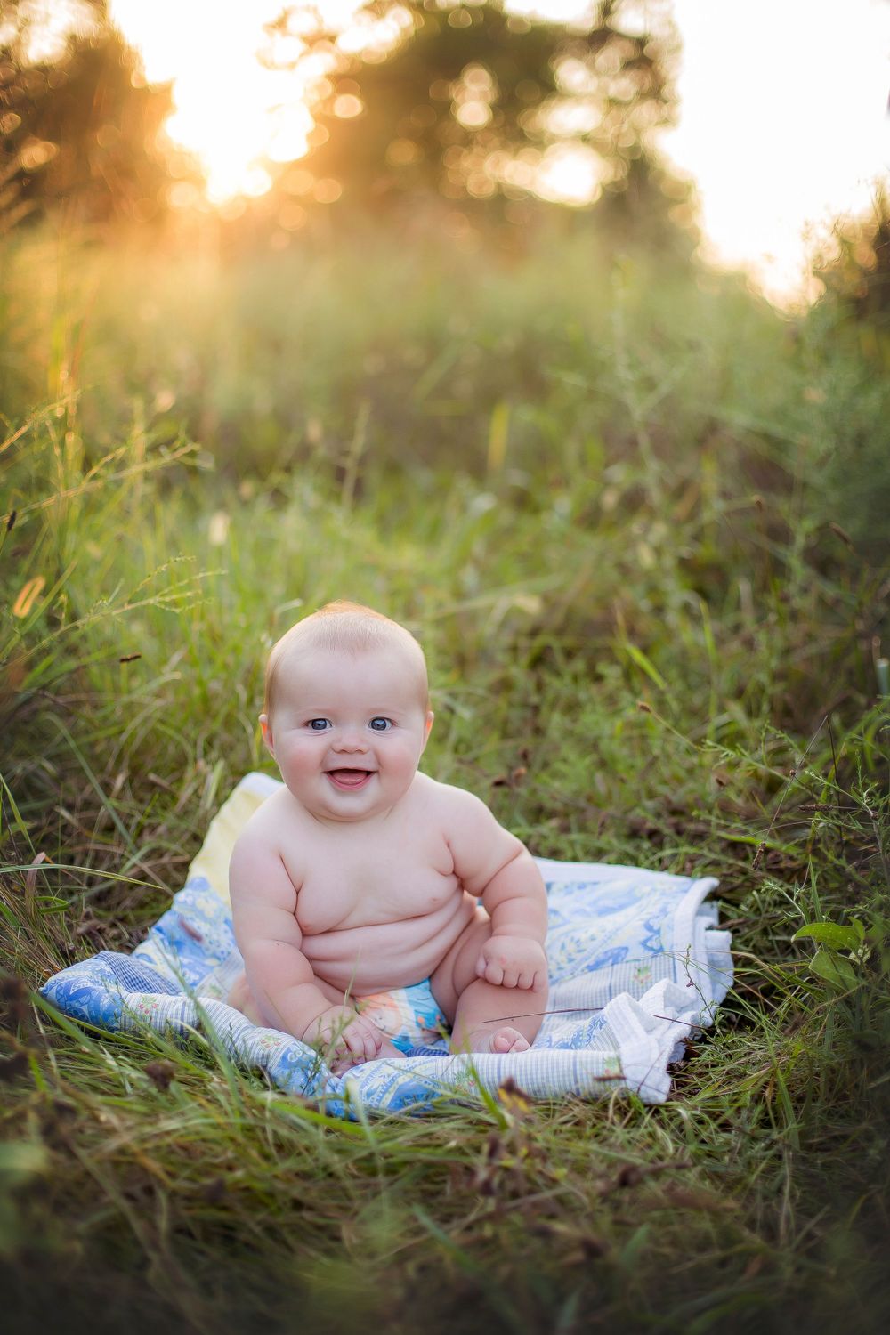 Six month old baby boy sitting in green field at sunset with blue and white blanket.  Baby is laughing.