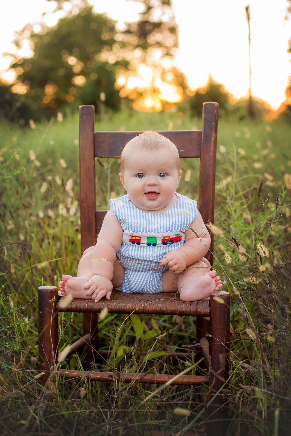 Six month old baby boy sitting on a chair in a field at sunset.  Baby is wearing a blue and white striped romper.