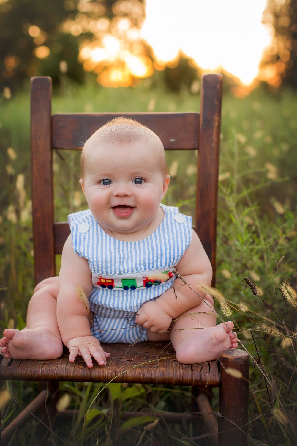 Six month old baby boy sitting on a chair in a field smiling at the camera.