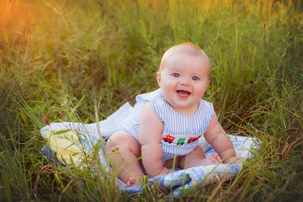 Six month old baby boy in a field sitting on a blanket.  He is leaning to the right side of the image laughing.