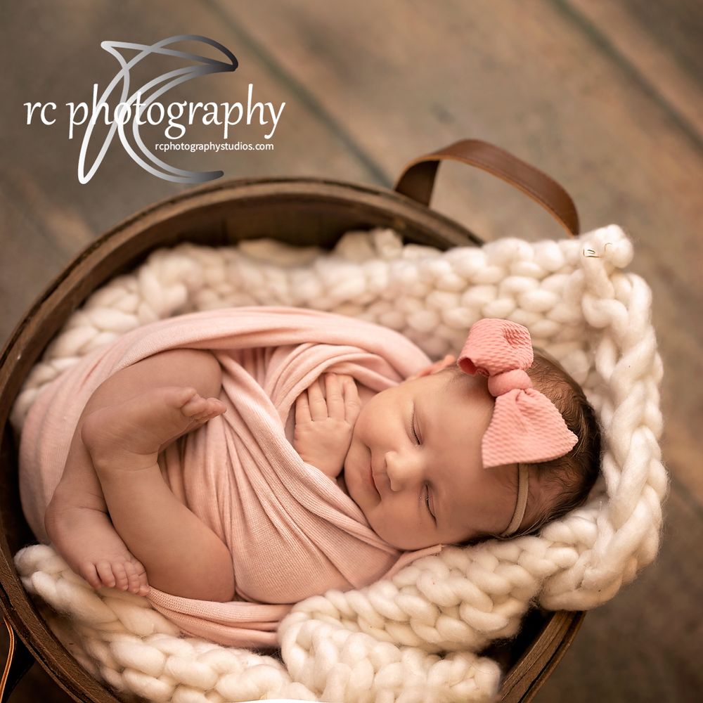 Peaceful newborn wrapped in pink swaddle sleeping in brown basket with knit blanket.