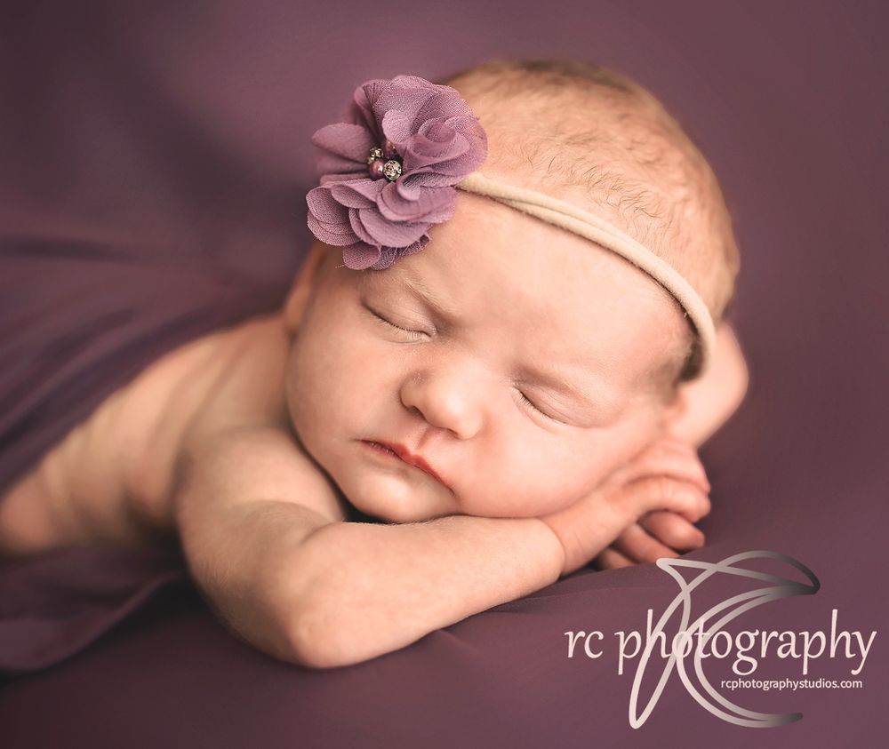Sleeping newborn wearing purple flower headband on purple background.