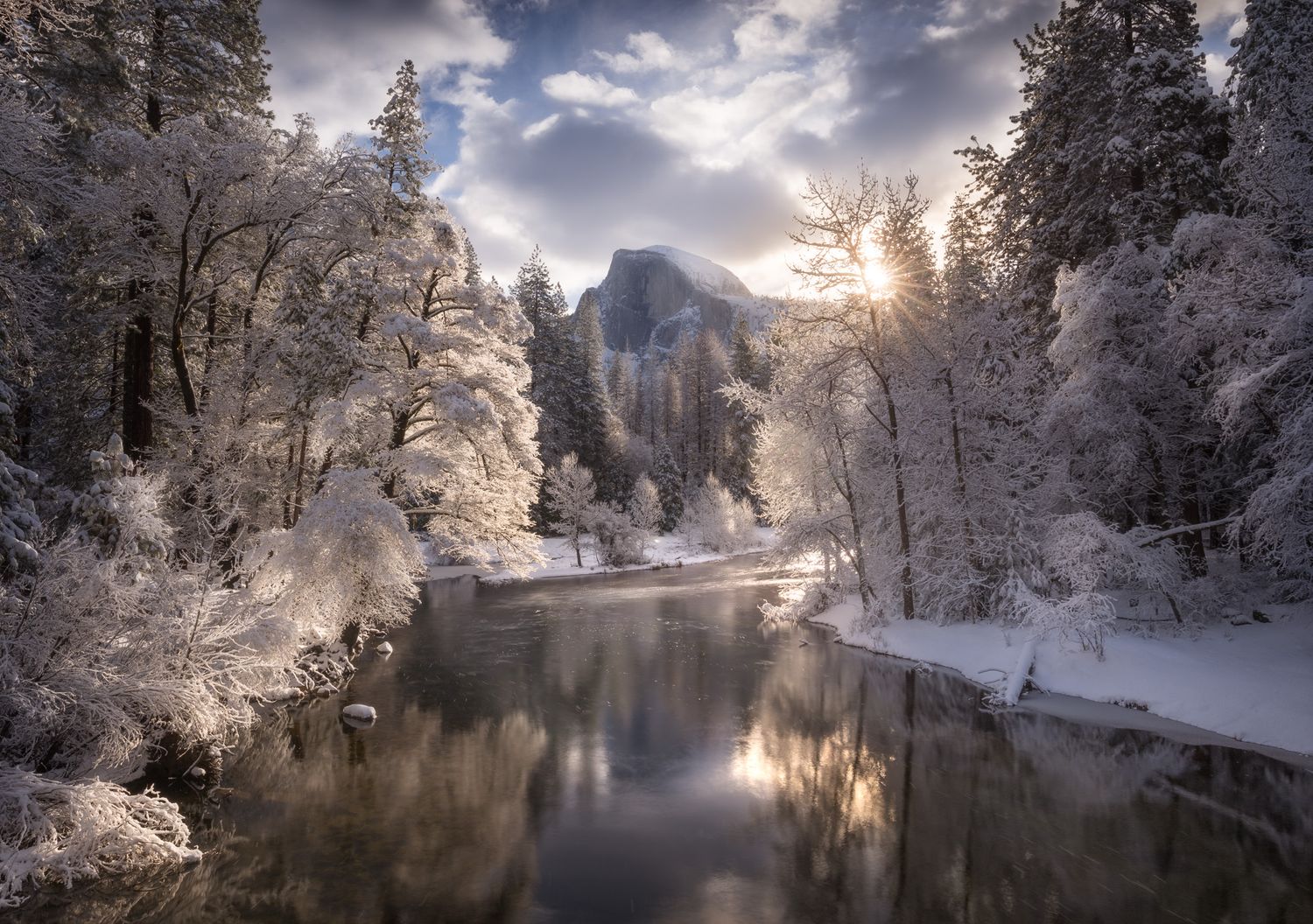 Snowy winter landscape in Yosemite Valley with Half Dome mountain reflected in Merced River at sunset.