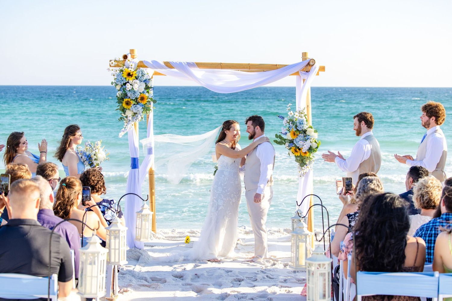 Beach wedding ceremony setup with floral arch and guests watching couple exchange vows by the ocean.