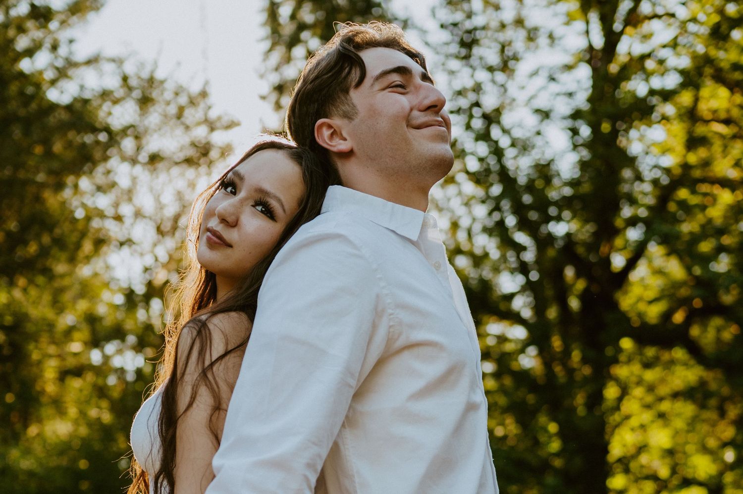 A couple laughs joyfully together in a sun-dappled outdoor setting with trees in the background.