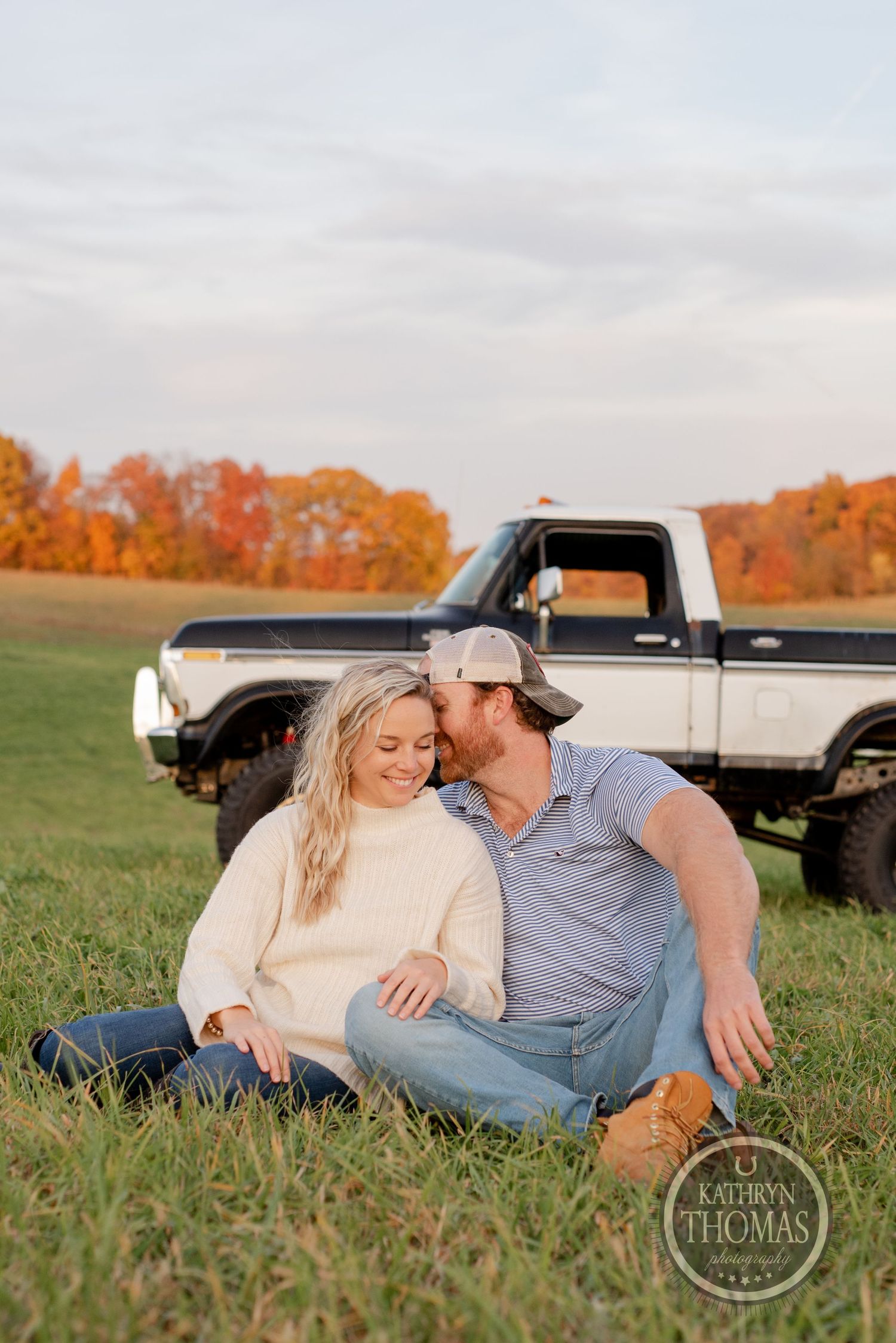 A couple sitting in a grassy field with a vintage pickup truck and autumn foliage in the background.