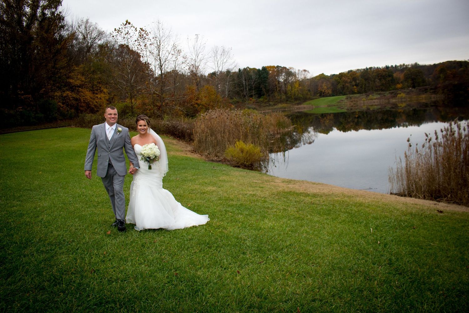 Wedding couple walking on green lawn beside autumn pond on overcast day.