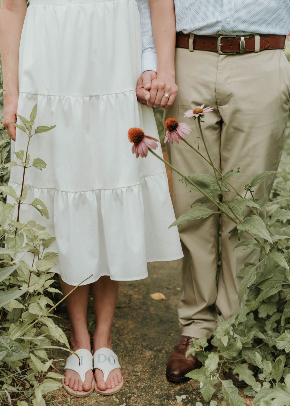 Detail shot of couple holding hands among wildflowers, showing white dress and khaki pants.