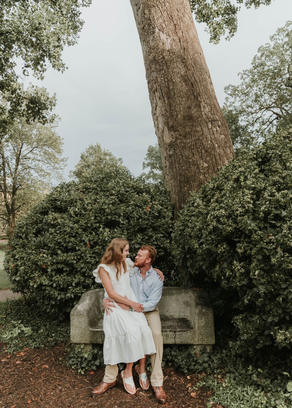 A couple sits together on a concrete ledge beneath a large tree in a garden setting.