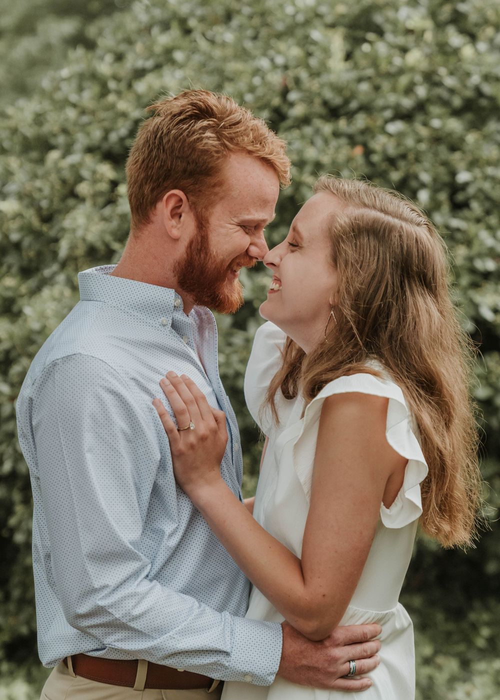 A couple smiles at each other in an intimate pose against green foliage.