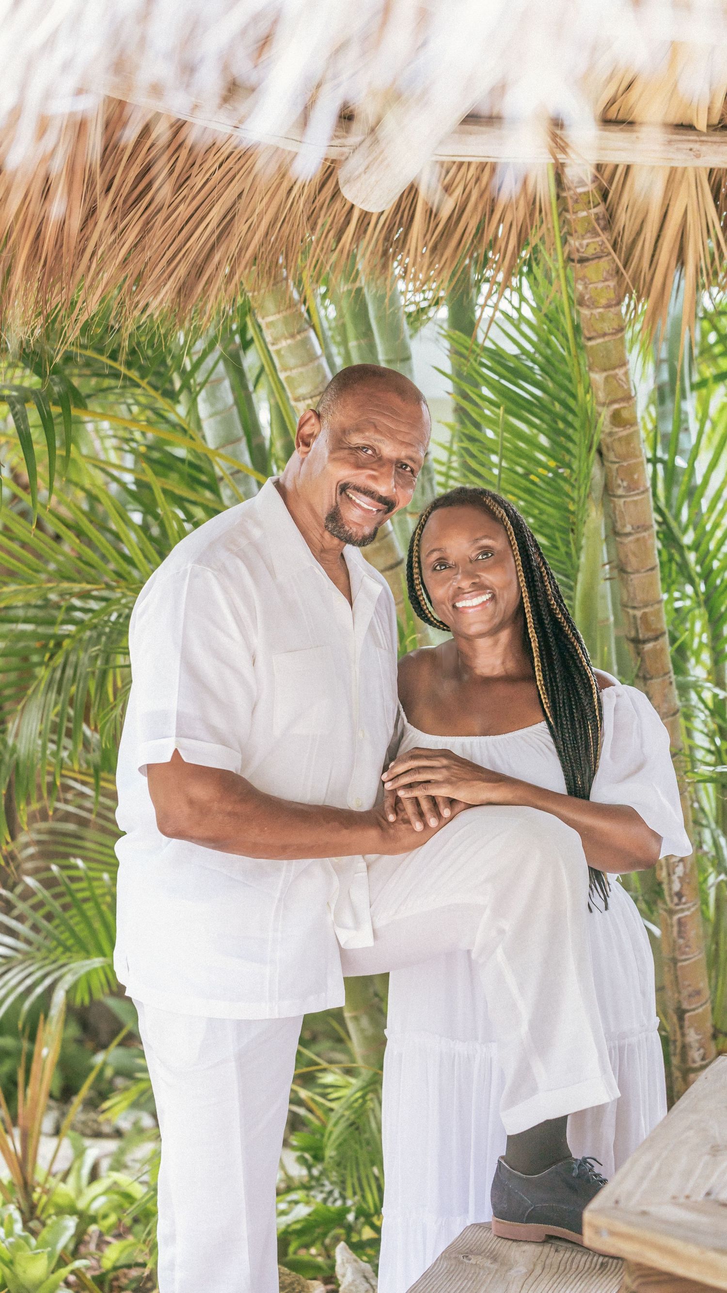 A couple wearing matching white outfits embraces while standing among tropical palm trees at a beachside location.