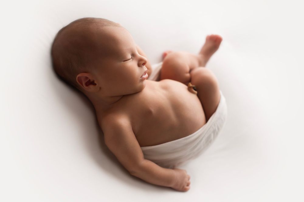 Backlit image of newborn baby boy laying on his back wrapped in a white wrap.
