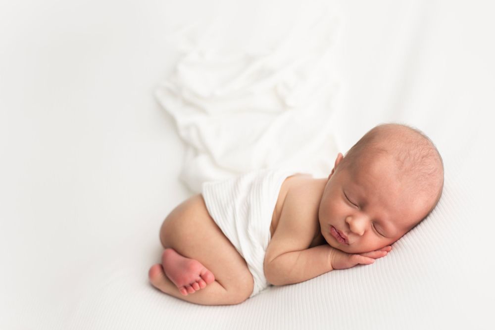 Newborn baby boy laying on white backdrop with white wrap, wrapped around baby's belly.