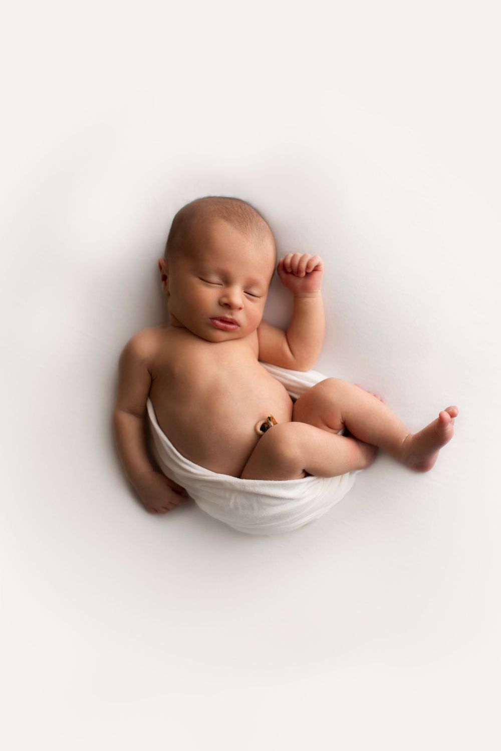 Newborn baby boy laying on back with white backdrop and white wrap around his bottom, curled into cozy position.