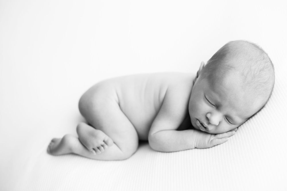 Black and white image with newborn baby boy laying on tummy, white backdrop with hand under his cheek.