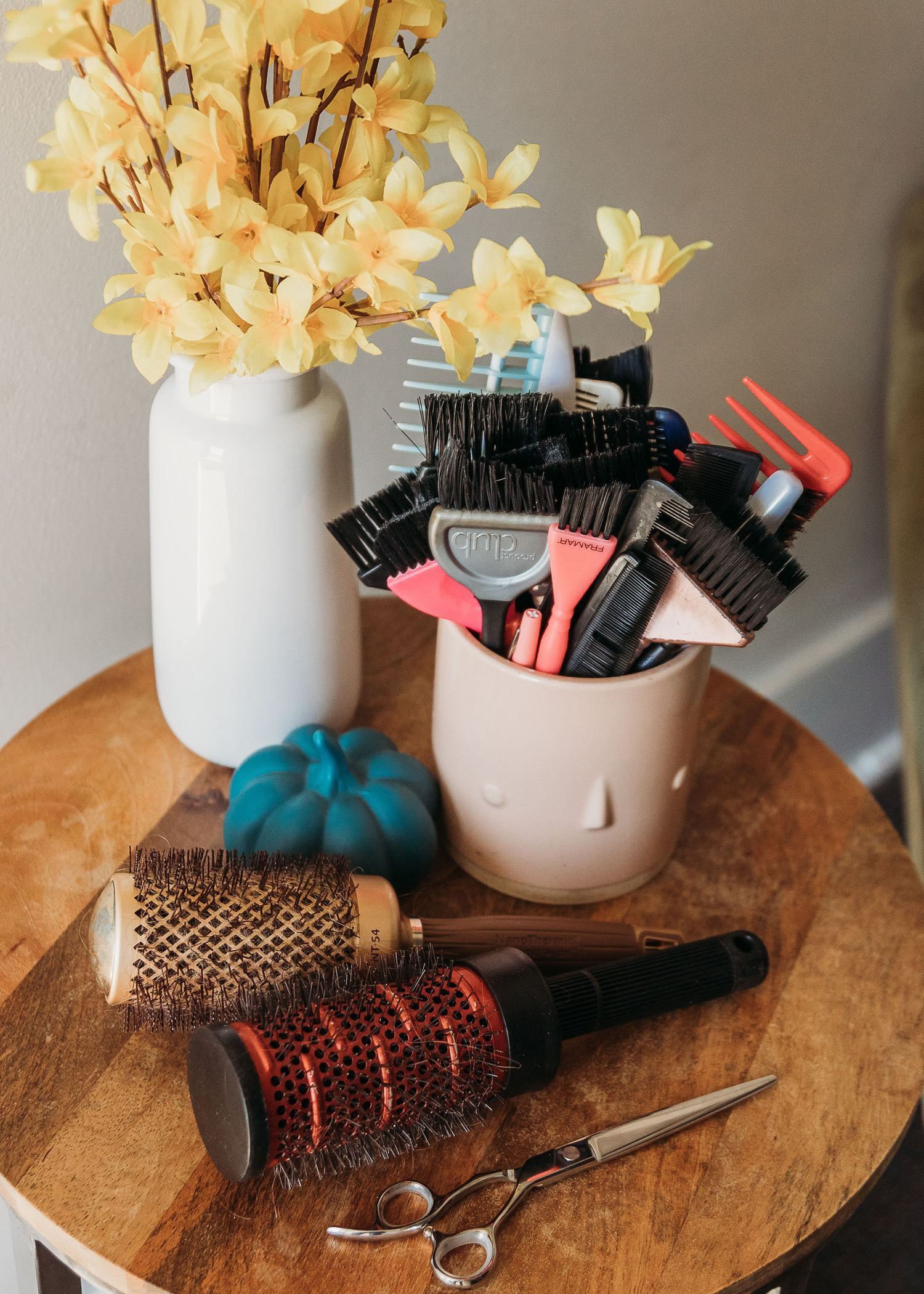 Hair styling tools and yellow flowers arranged on wooden surface with teal pumpkin accent.