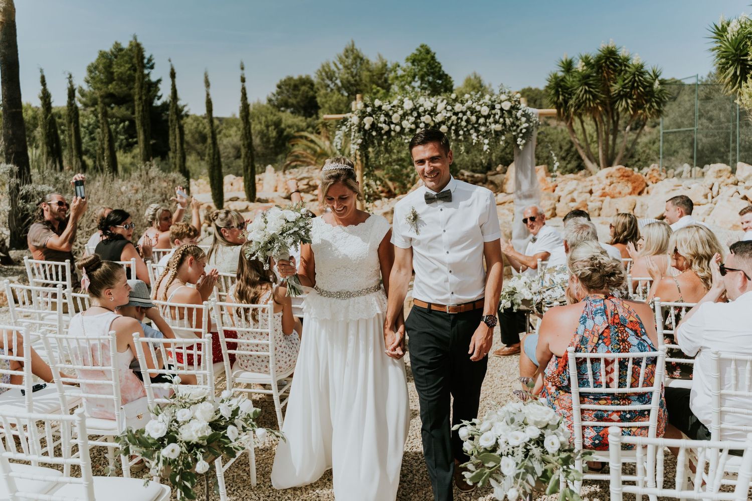 Outdoor wedding ceremony with white chairs, floral decorations and cypress trees creating a romantic atmosphere.