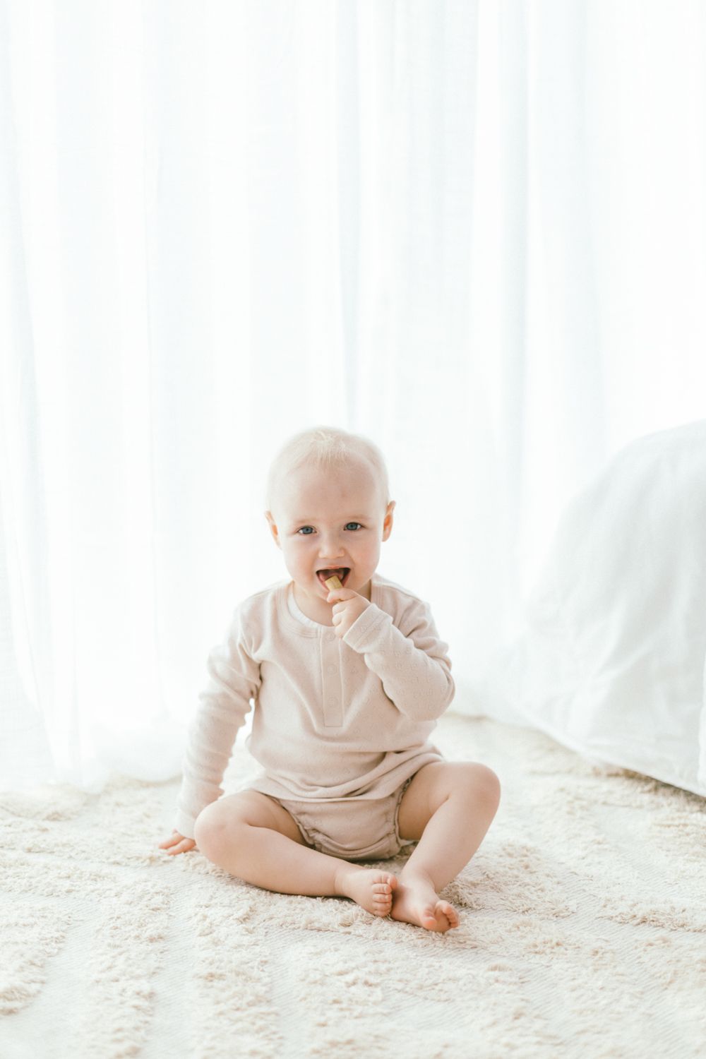 Baby in cream-colored onesie sits on plush white carpet in bright, minimalist setting.