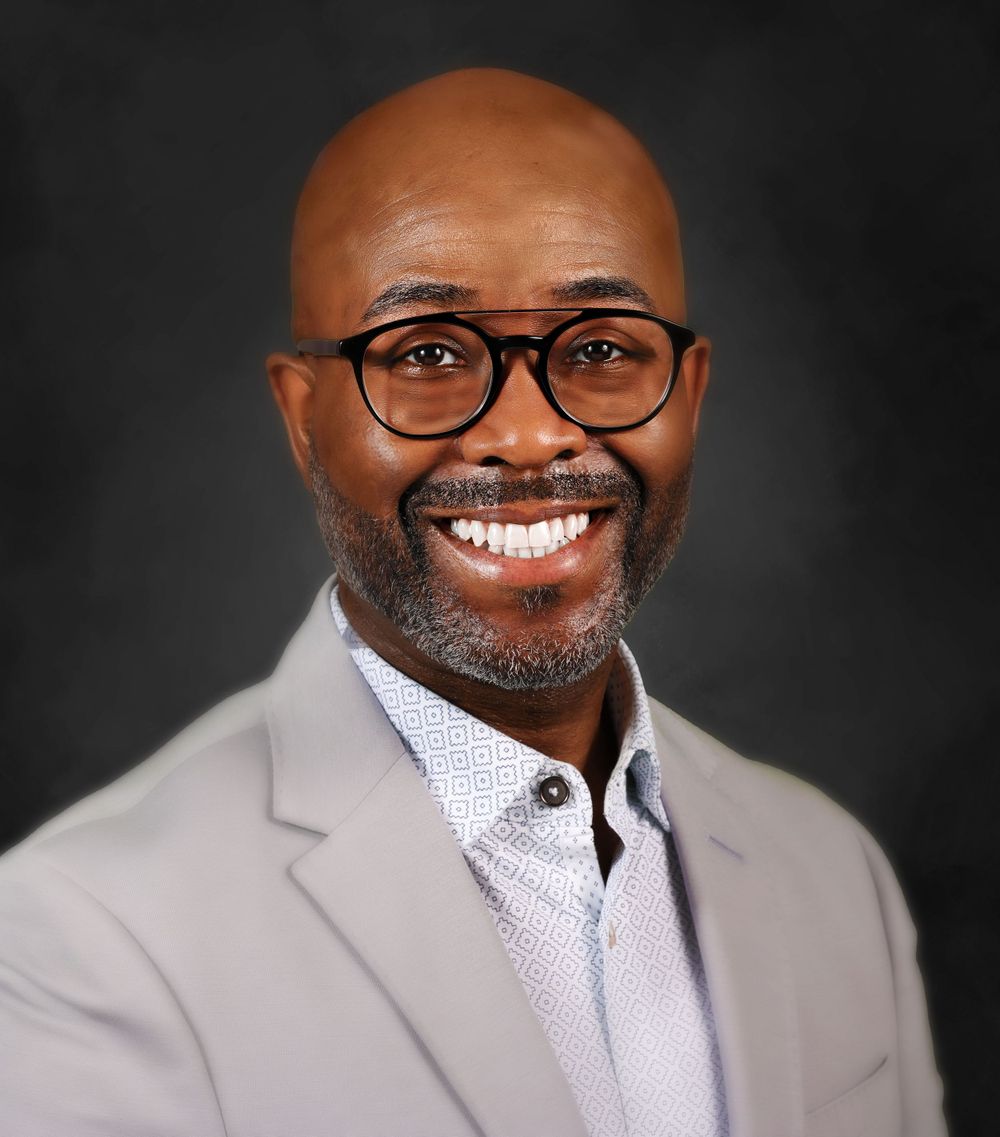Professional headshot of person in grey blazer and glasses against dark background.