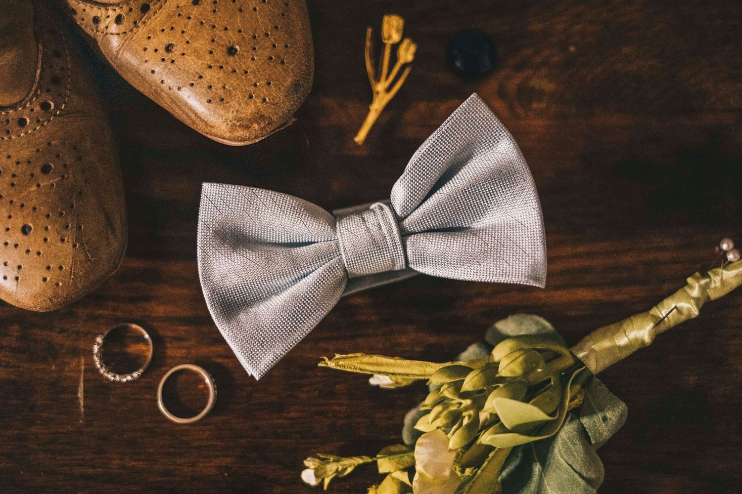 Gray bow tie with brown brogue shoes, gold rings, and floral boutonniere on rustic wooden table.