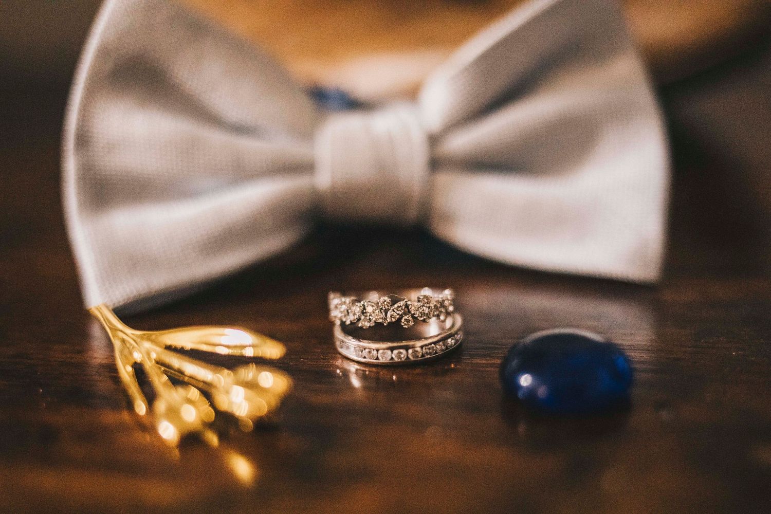Diamond crown wedding ring and blue sapphire cufflink beside gold boutonniere and gray bow tie.