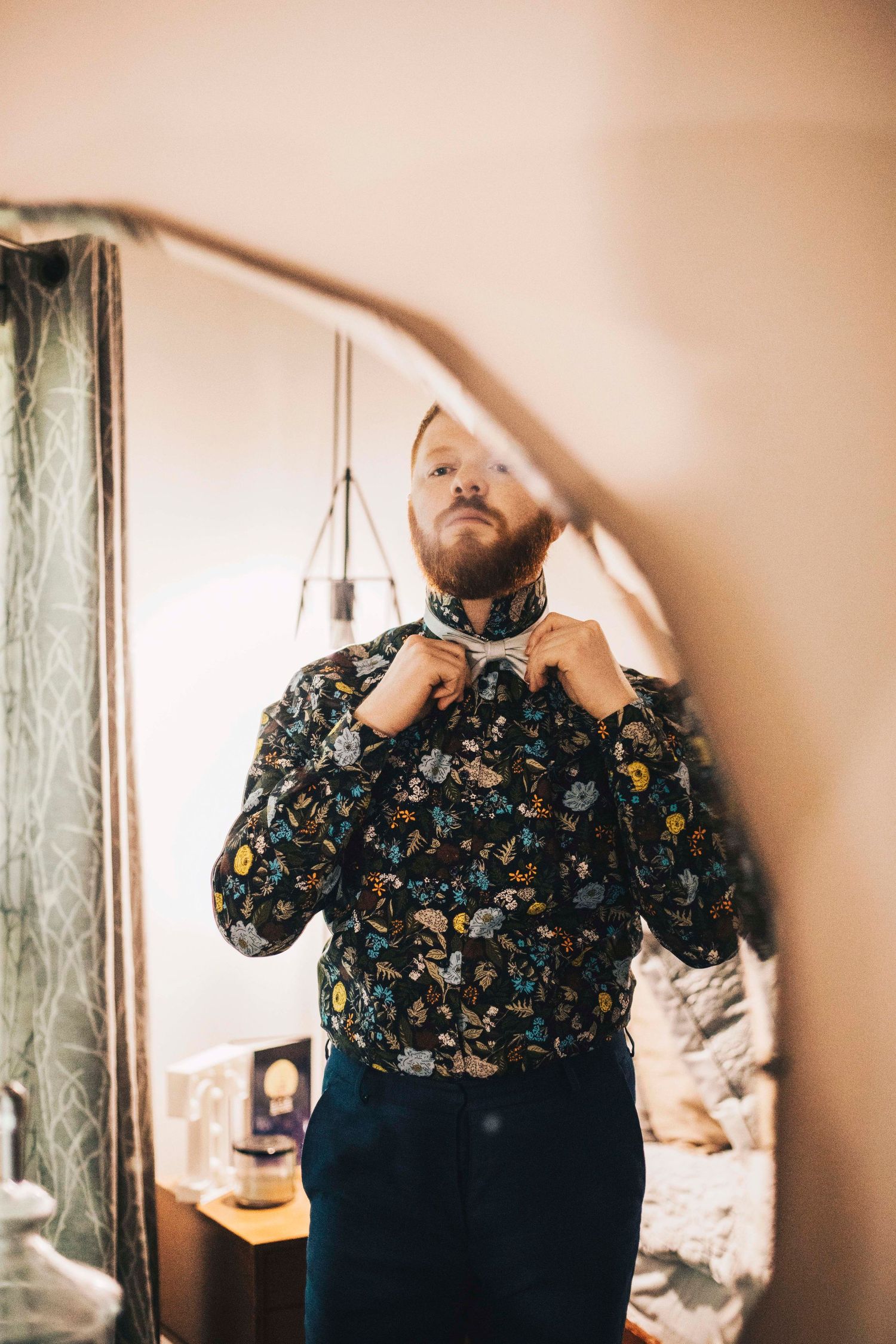 Bearded groom in floral shirt adjusting bow tie while looking in mirror before wedding.