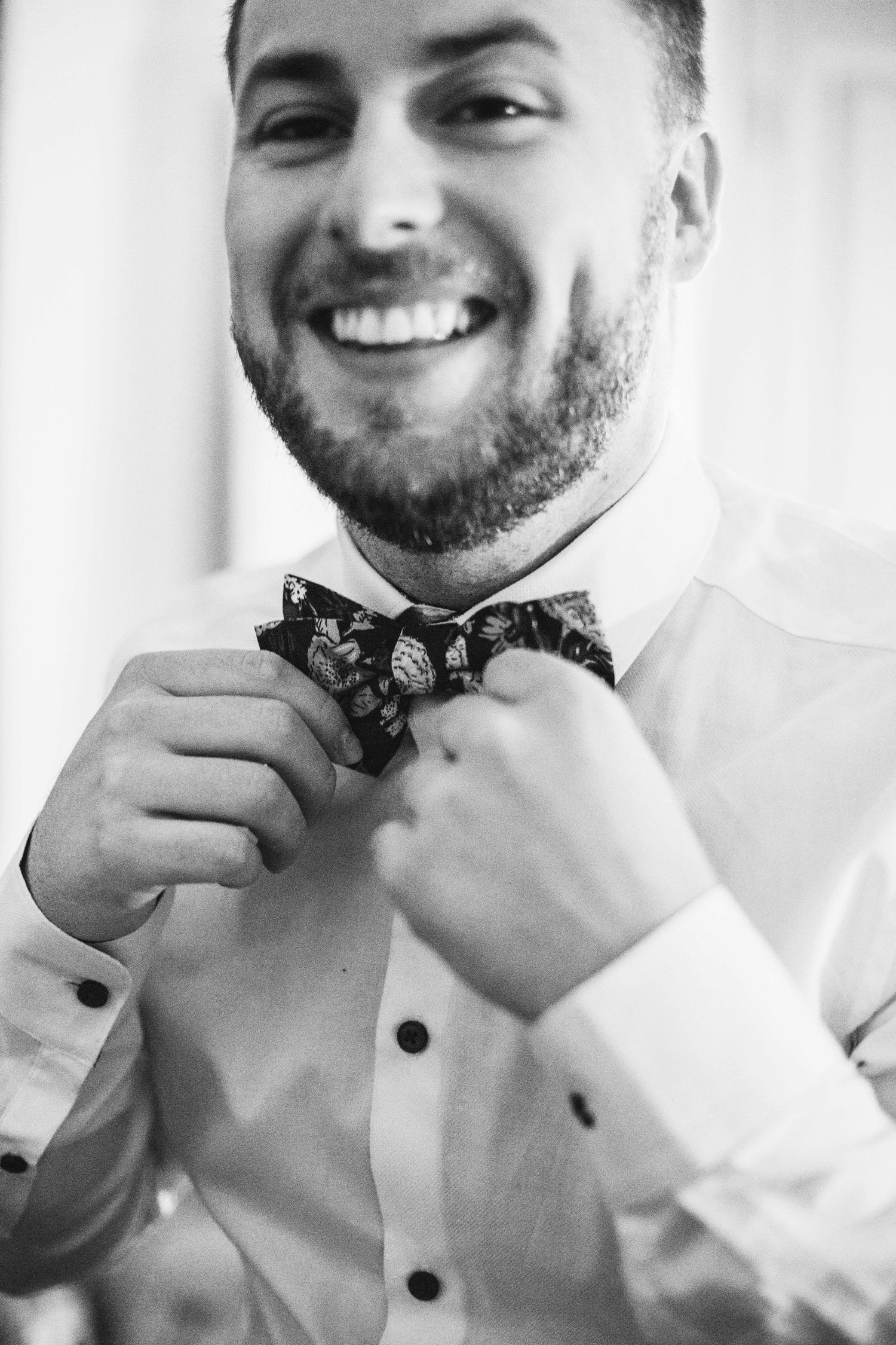Smiling groom in white shirt adjusting patterned bow tie in black and white wedding photo.