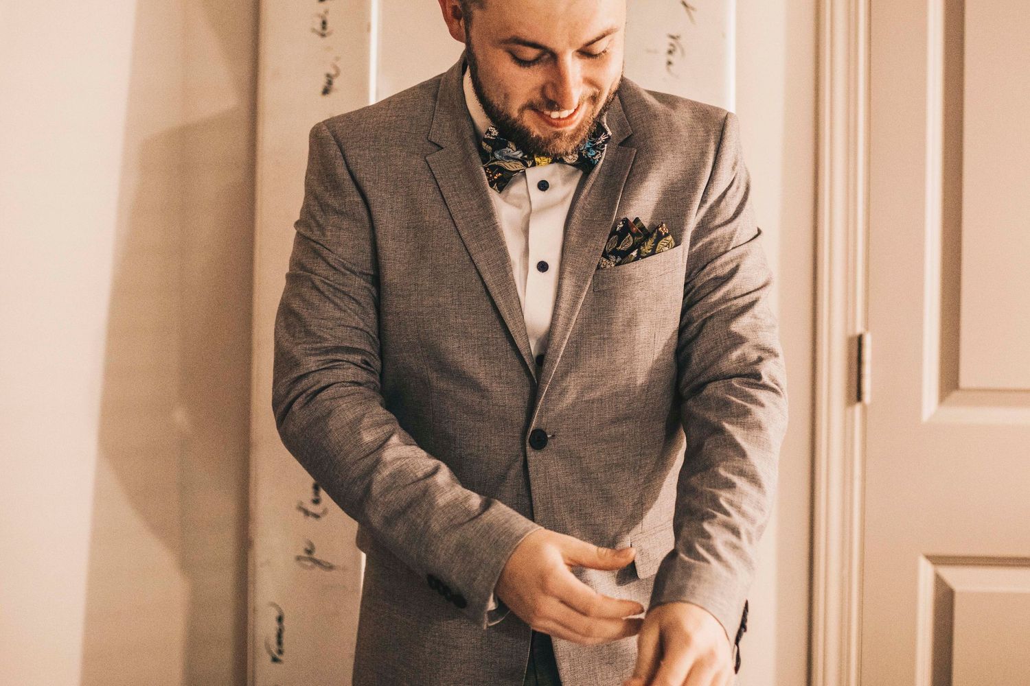 Groom in gray suit with patterned bow tie and pocket square adjusting cufflinks before ceremony.