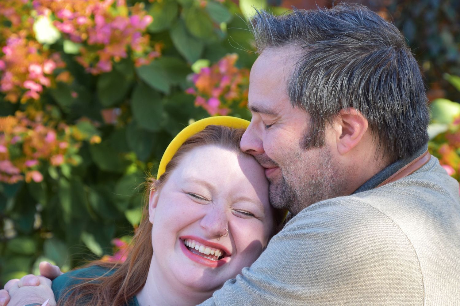 A couple laughs joyfully together outdoors, with the man embracing and kissing the red-haired woman near pink flowers.