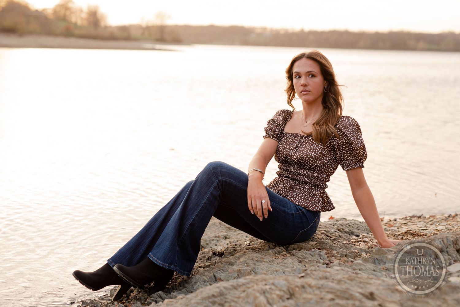 Someone in a leopard print top and flare jeans poses on a rocky shoreline at golden hour.