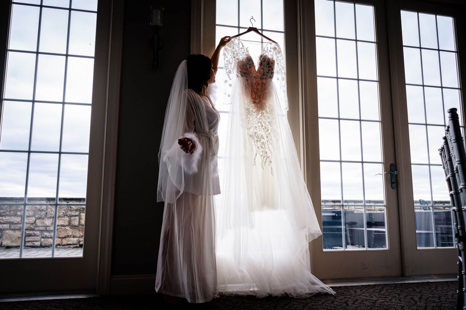 Wedding dress and veil displayed in front of large industrial windows with dramatic lighting.