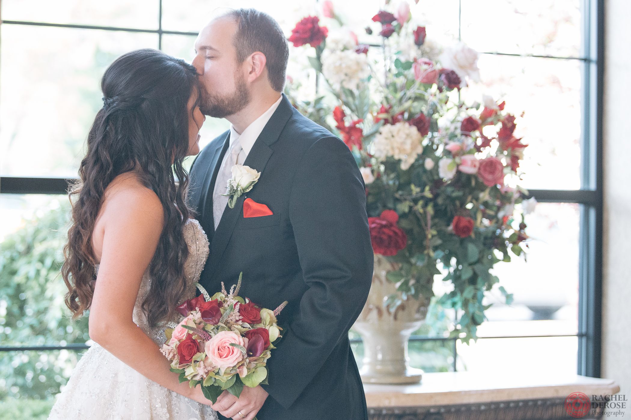 A wedding couple sharing an intimate moment with red and white floral arrangements in the background.