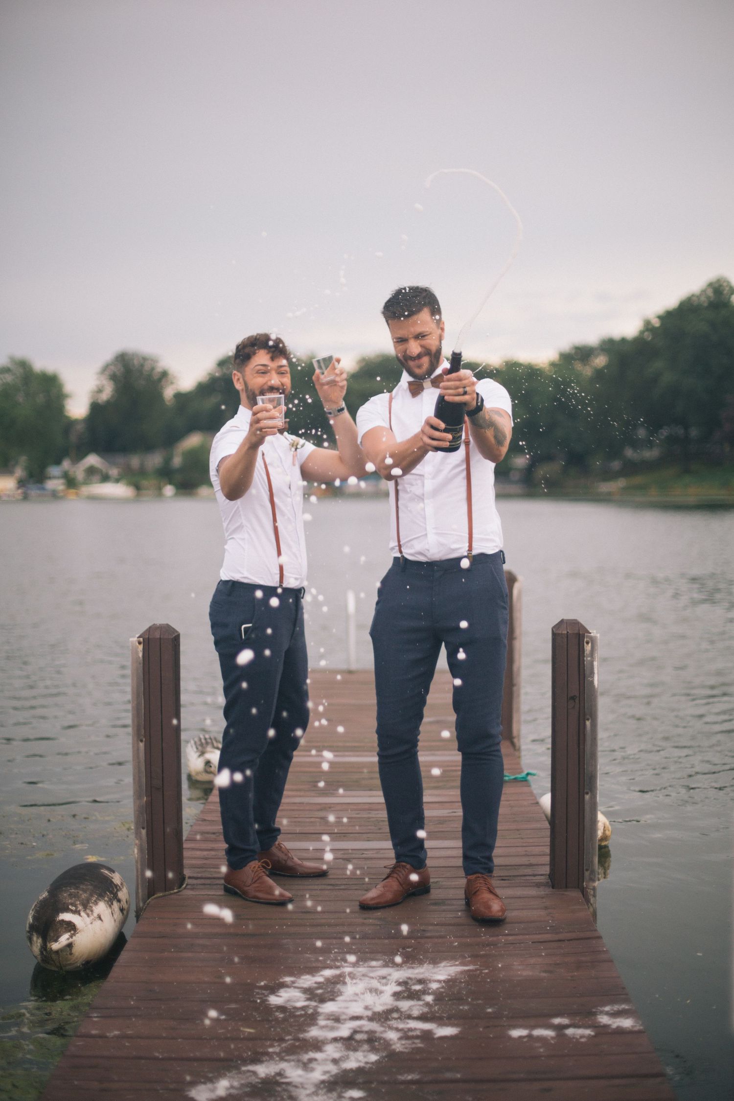 Two people in suspenders celebrate with champagne on a wooden dock by the lake.