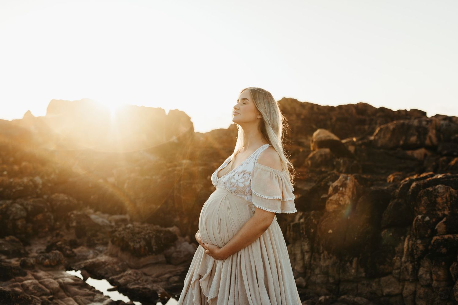 A pregnant woman in a white dress stands against a rocky landscape during golden hour sunset.