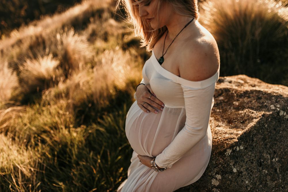 Silhouette of expectant mother in white dress against golden sunset on mountainside.