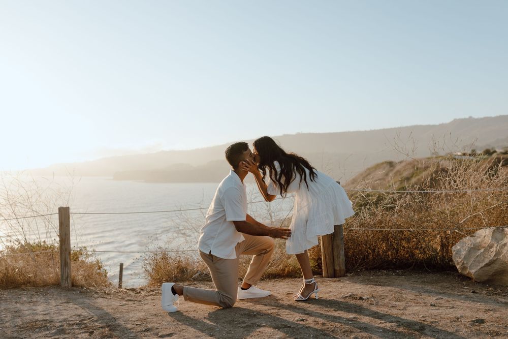Photo of a proposal. Man is on one knee, while the woman bends down to kiss him. Cliff & ocean backdrop. Golden Hour.