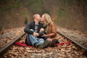 A romantic fall scene with a couple sitting on train tracks surrounded by autumn leaves and a red blanket.