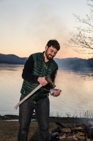 Person chopping wood by a lakeside at sunset with mountains in the background.