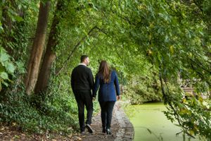 Two people walk along a shaded path beside a tranquil green pond surrounded by lush foliage.