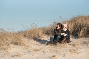 A couple sits together on sandy dunes with tall grasses swaying in the breeze against a blue sky.