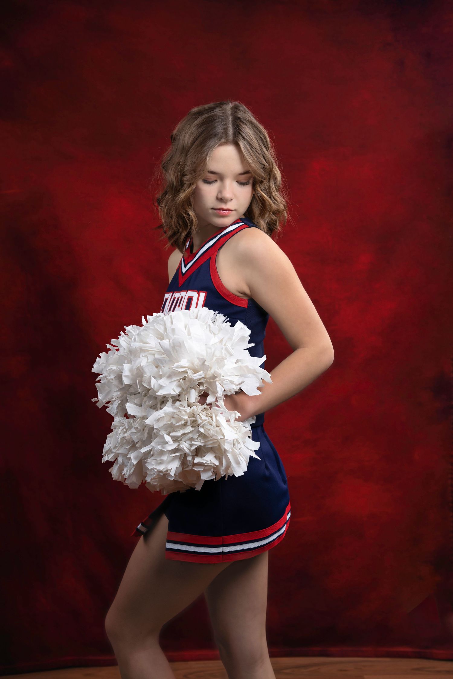 Cheerleader in navy uniform poses with white pom poms against red dramatic backdrop.