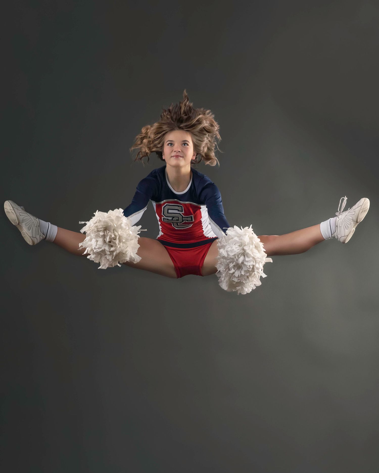 Cheerleader performs toe touch jump with pom poms against dark studio background.