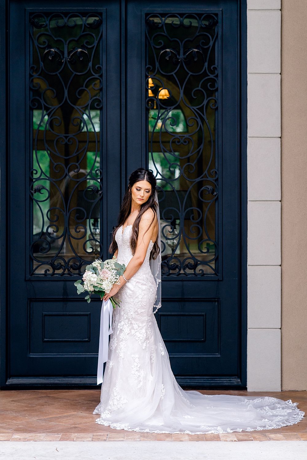 Bride in lace wedding gown standing before ornate black doors holding a white and green bouquet.