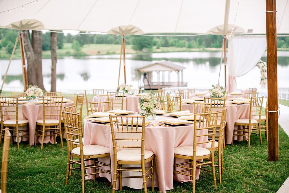Elegant outdoor wedding reception setup with pink tablecloths and gold chiavari chairs beside a lake.