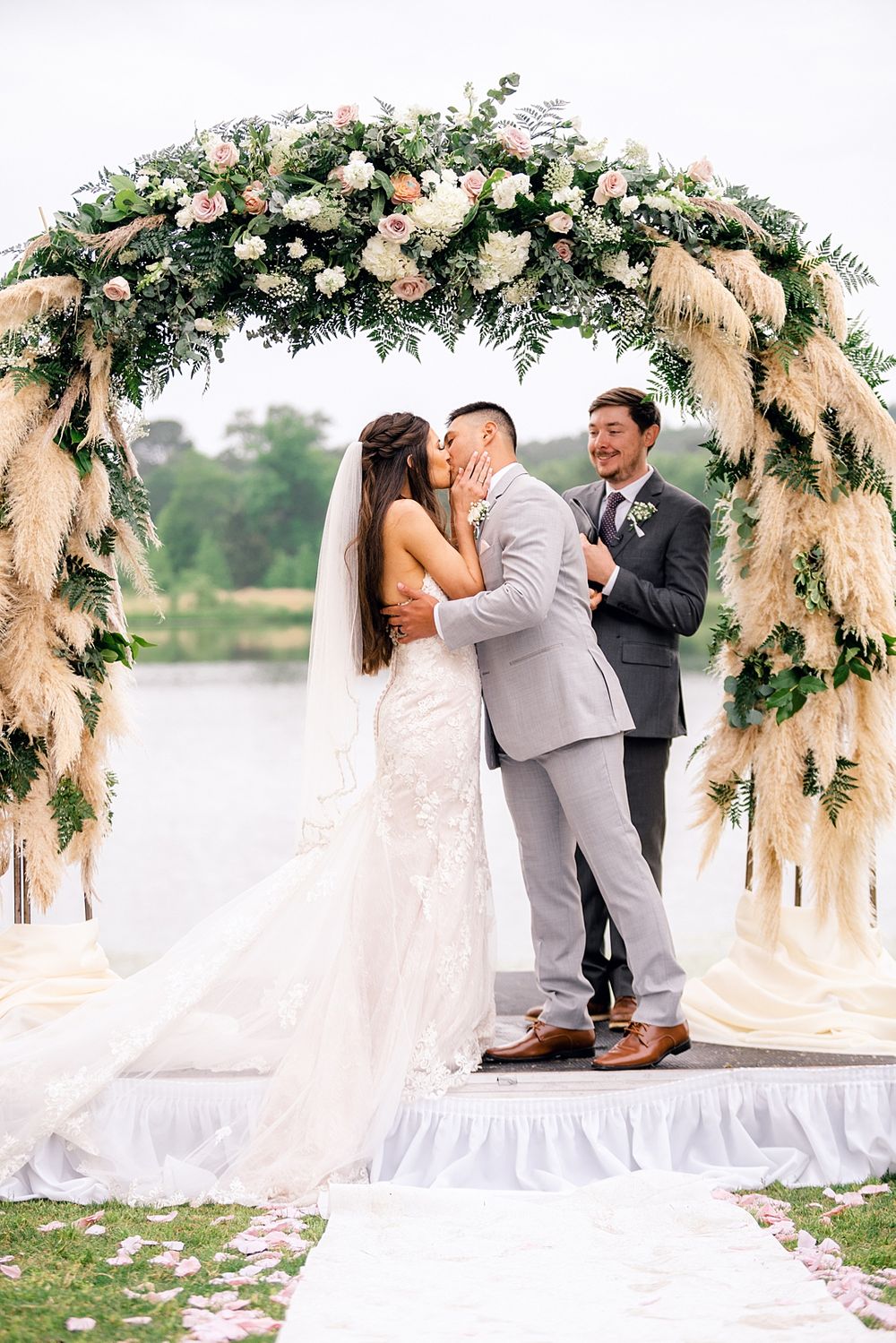 Wedding ceremony kiss under an ornate arch decorated with roses and pampas grass.
