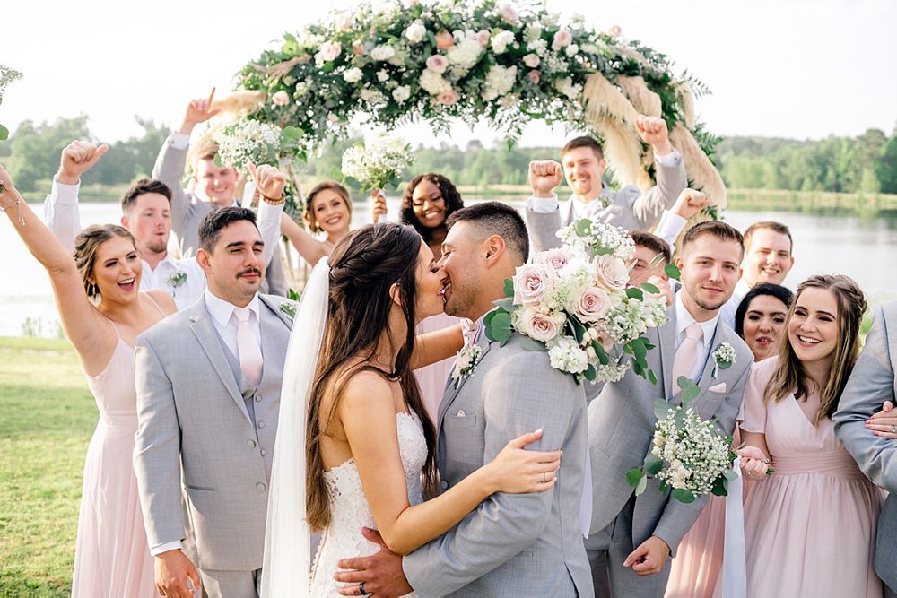 Wedding party celebrating under a floral arch with pink roses and white blooms outdoors.