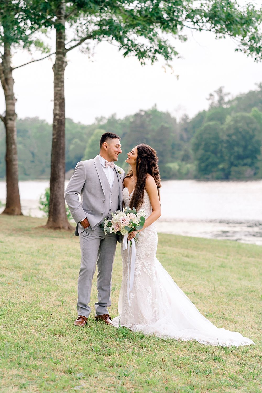 Lakeside wedding portrait featuring a couple in grey suit and lace gown beneath tall trees.
