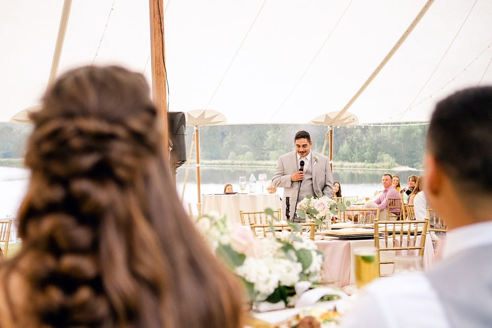 A wedding speech being delivered in an elegant outdoor tent reception setting with table settings and floral decor.